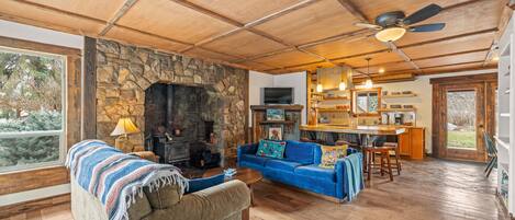 A cozy living room with a stone fireplace, a blue sofa, a wooden ceiling, and a kitchen area with a bar.