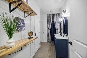 Un pasillo con un estante de madera, un jarrón blanco con plantas verdes, un reloj y un baño con lavamanos y espejo.