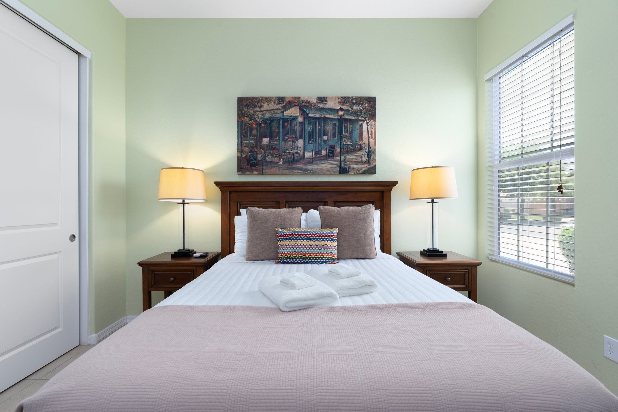 A neatly made bed with a colorful pillow, two bedside tables with lamps, a painting above the bed, and a window with blinds.