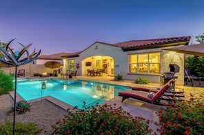 Zona de piscina bien iluminada con sillones de descanso, un patio y una casa con fachada de piedra y grandes ventanales.