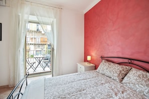 Une chambre à coucher avec un lit, une table de chevet, une lampe, une fenêtre avec des rideaux et une vue sur les bâtiments à l’extérieur.