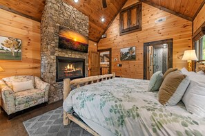 A cozy log cabin bedroom with a stone fireplace, a bed with floral bedding, a wooden bench, and a chair.