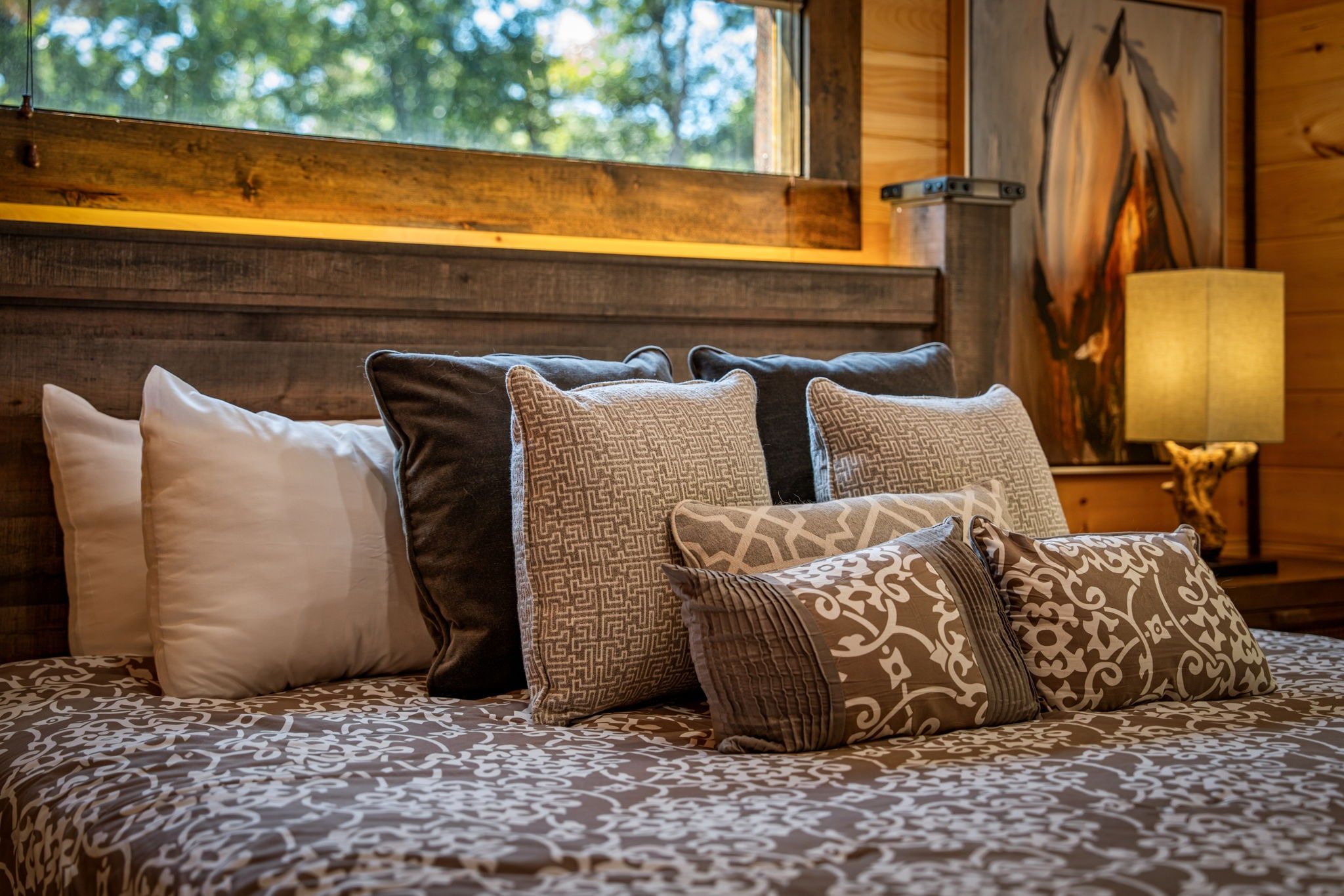 Cama com várias almofadas e colcha estampada em quarto com paredes de madeira e janela.