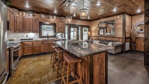 A spacious kitchen with wooden cabinets, a large island with stools, and a stone countertop. - Jazzy's Cozy Creekside - Hot Tub | Game Room | Screened Decks (Blue Ridge)