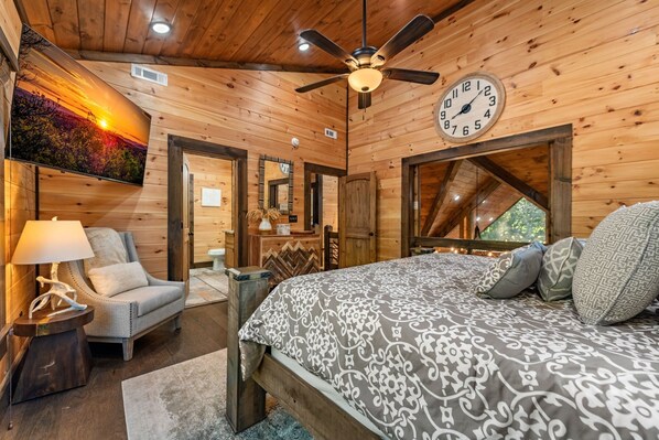 Interior de una cabaña de madera con una cama, una silla, un ventilador de techo, un reloj y un cuadro.