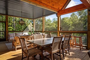 A wooden deck with a dining table and chairs, surrounded by large windows offering a view of trees.