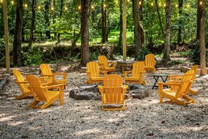 An outdoor seating area with Adirondack chairs and a table surrounded by rocks, set in a forested area.