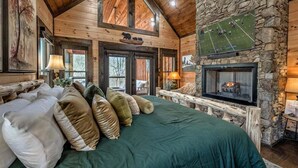 A cozy log cabin bedroom with a large bed, a stone fireplace, and a mounted sports game. - Jazzy's Cozy Creekside - Hot Tub | Game Room | Screened Decks (Blue Ridge)