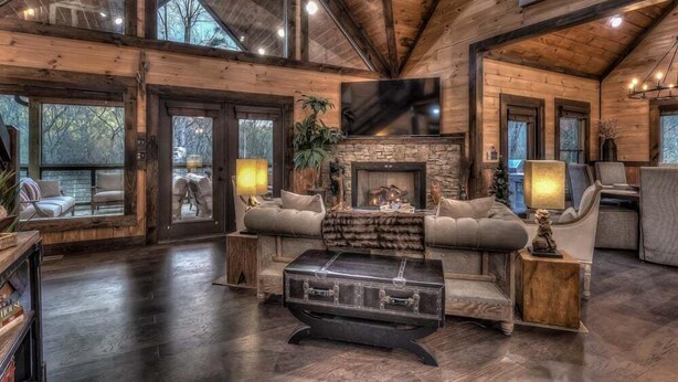 A spacious living room with a stone fireplace, comfortable seating, and a large flat-screen TV mounted on the wall. - Jazzy's Cozy Creekside - Hot Tub | Game Room | Screened Decks (Blue Ridge)
