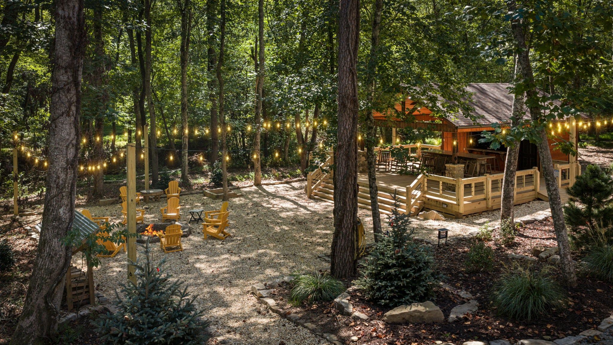 A forested area with a wooden cabin, outdoor seating, and string lights.