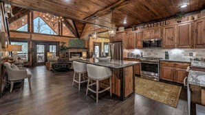 A spacious kitchen with wooden cabinets, a large island with bar stools, and a stone backsplash. - Jazzy's Cozy Creekside - Hot Tub | Game Room | Screened Decks (Blue Ridge)