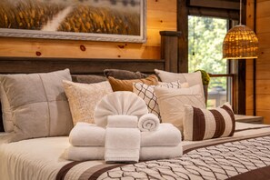 Een net opgemaakt bed met meerdere kussens en opgerolde handdoeken, een beddengoed met een patroon en een hangende lamp.
