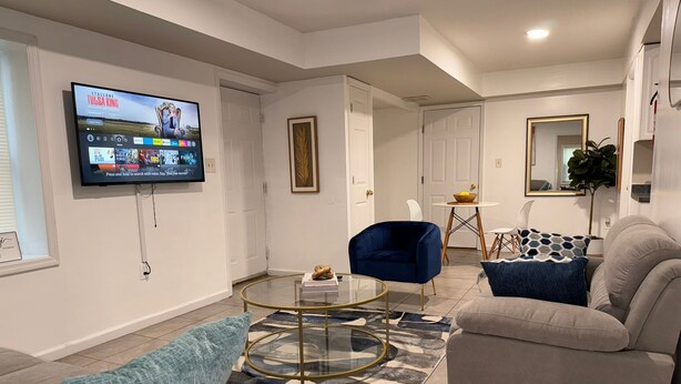 Ein modernes Wohnzimmer mit einem an der Wand montierten Flachbildfernseher, einem runden Glastisch, zwei Sofas mit blau-grauen Kissen und einem kleinen Essbereich mit Tisch und Stühlen.
