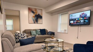 Ein modernes Wohnzimmer mit einem beigen Sofa, einem Glastisch und einem blauen Sessel. Ein Flachbildfernseher an der Wand zeigt einen Sportkanal.