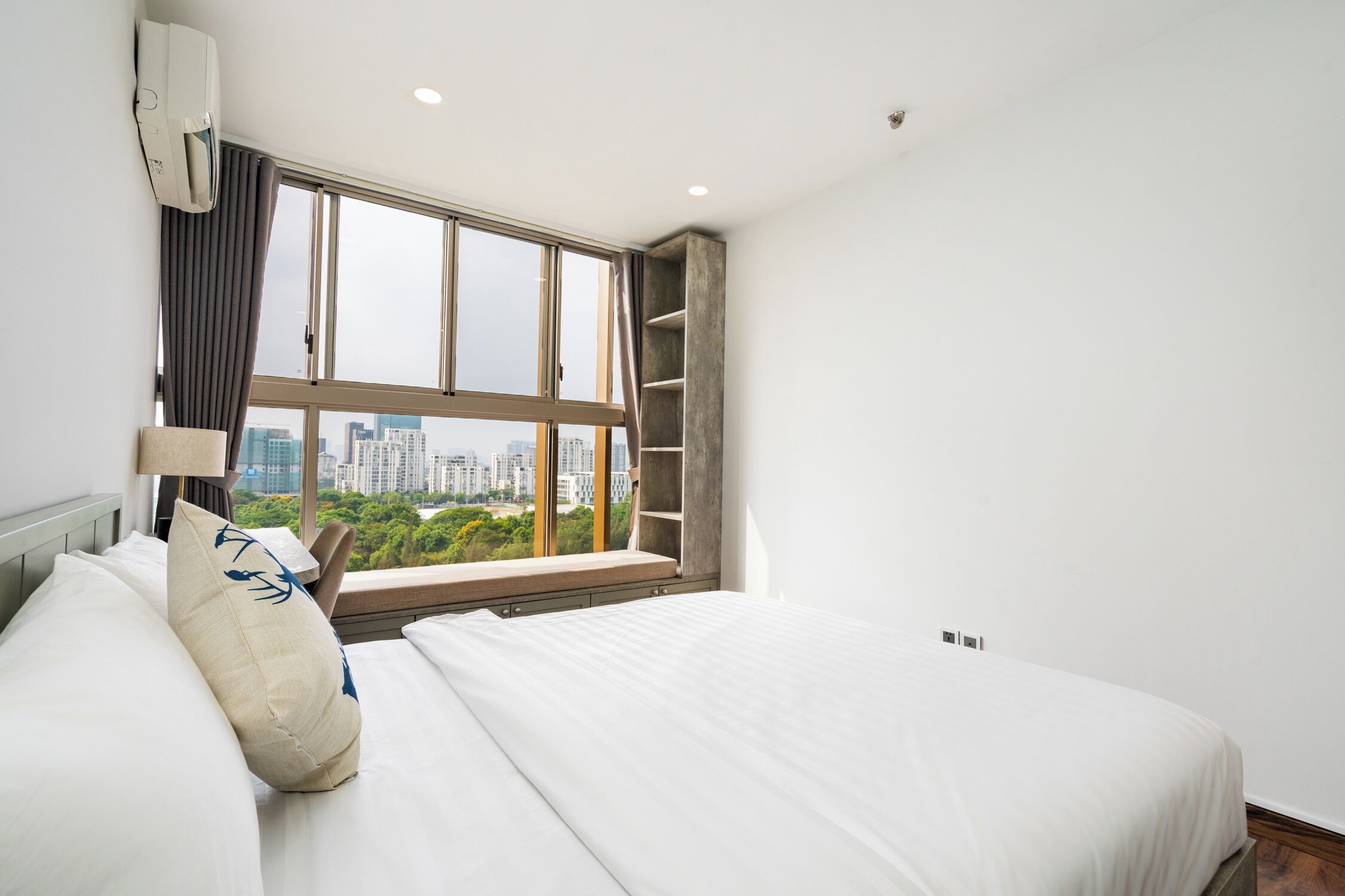 A modern hotel room with a large window, a bed with white linens, a built-in shelf, and a city view.
