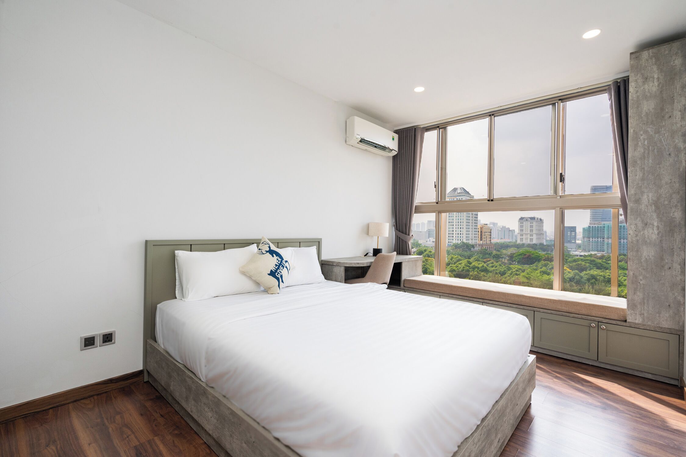 Habitación de hotel con una cama grande, un escritorio con una silla, un equipo de aire acondicionado y vistas de la ciudad a través de un ventanal amplio.