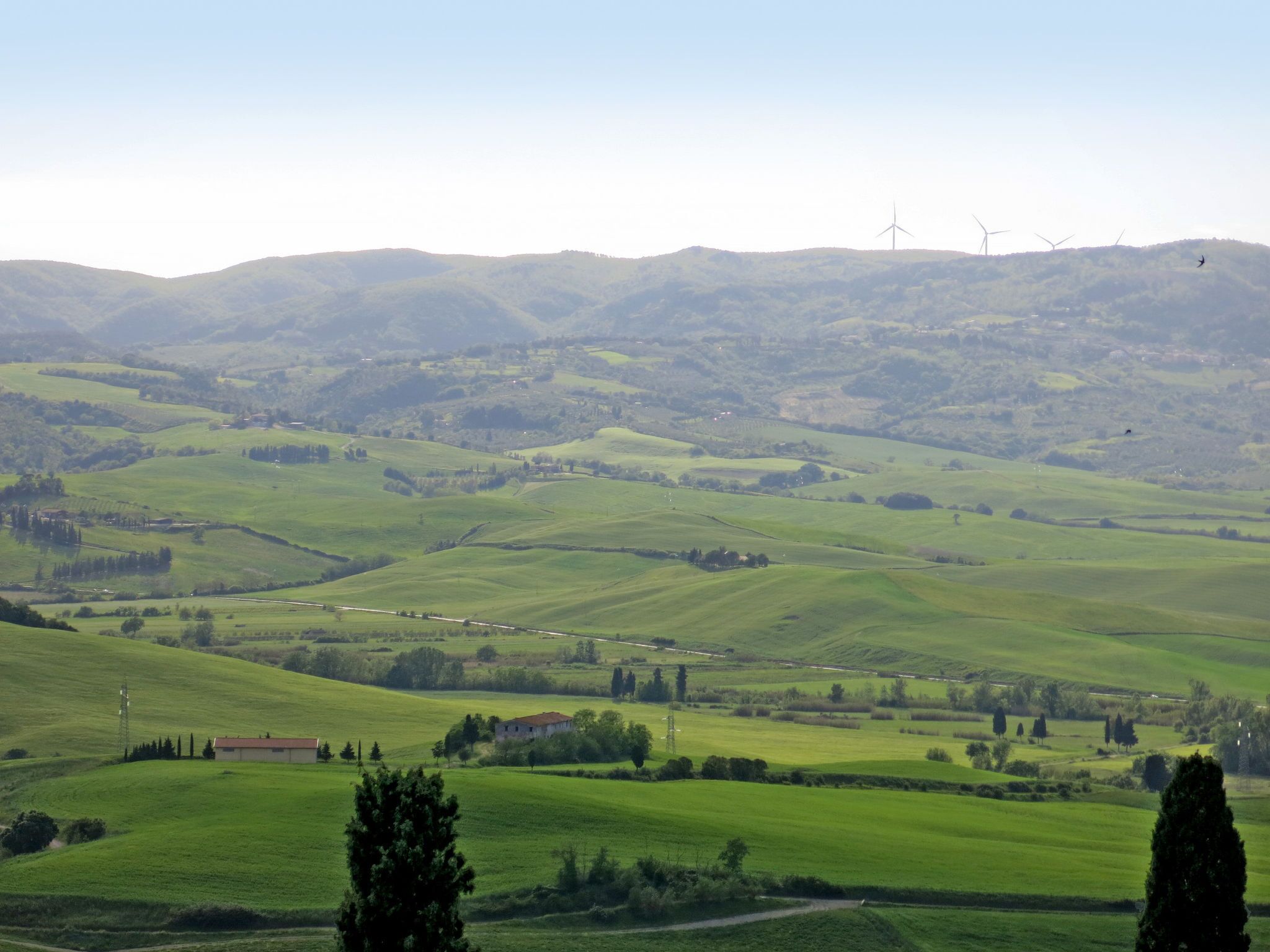 Paisaje rural con colinas ondulantes, algunos edificios y aerogeneradores a lo lejos.