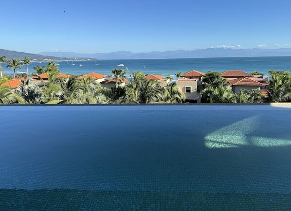 Vista deslumbrante de uma piscina infinita com paisagem costeira ao fundo, incluindo uma praia e montanhas distantes.