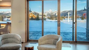 Ein modernes Interieur mit großen Schiebeglastüren, die zu einer verschneiten Außenanlage führen, mit einer Holzhütte und Bergen im Hintergrund.