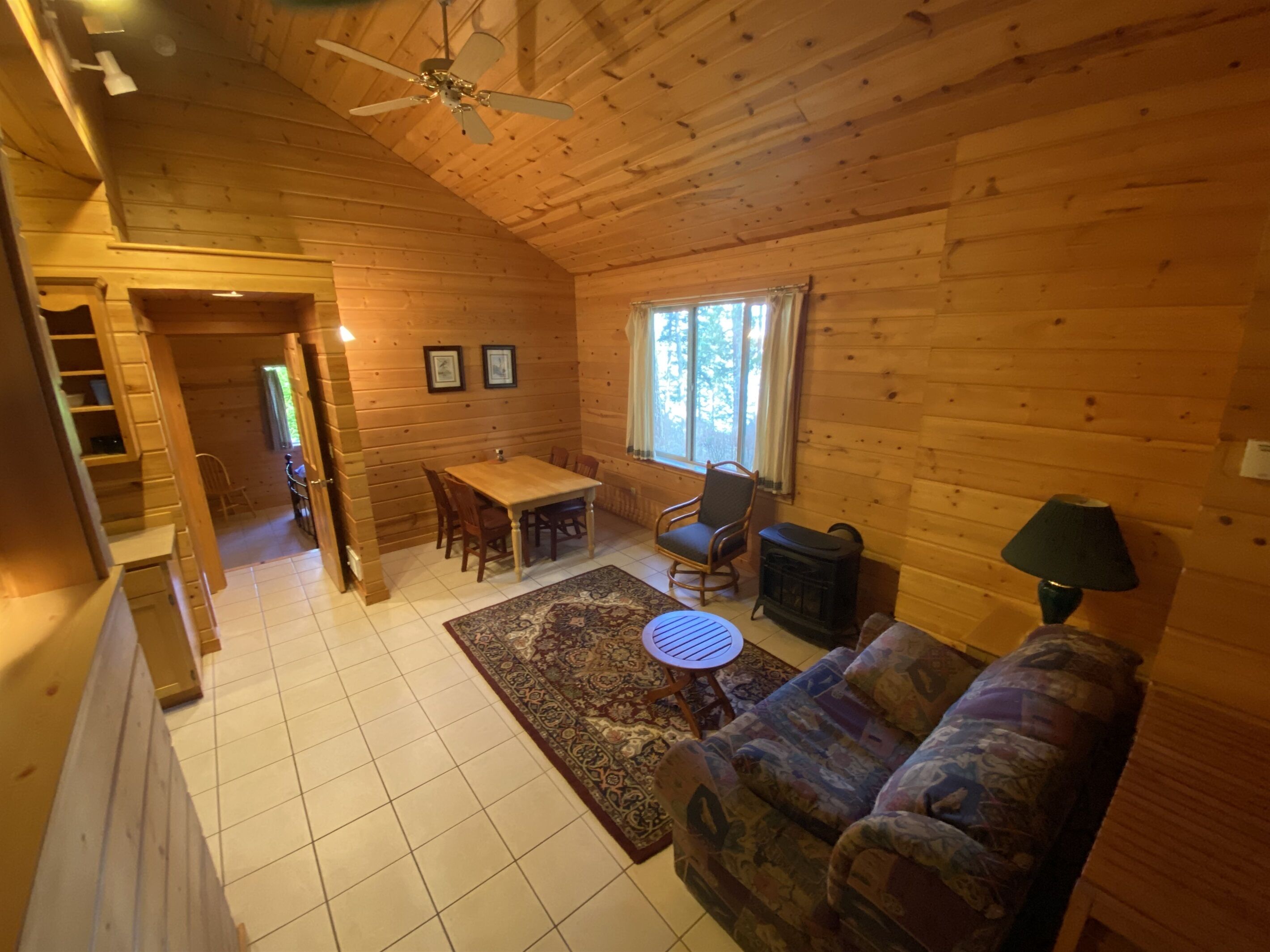 Een gezellig interieur van een chalet met een eethoek, een bank, een stoel, een klein tafeltje, een kachel en een plafondventilator.