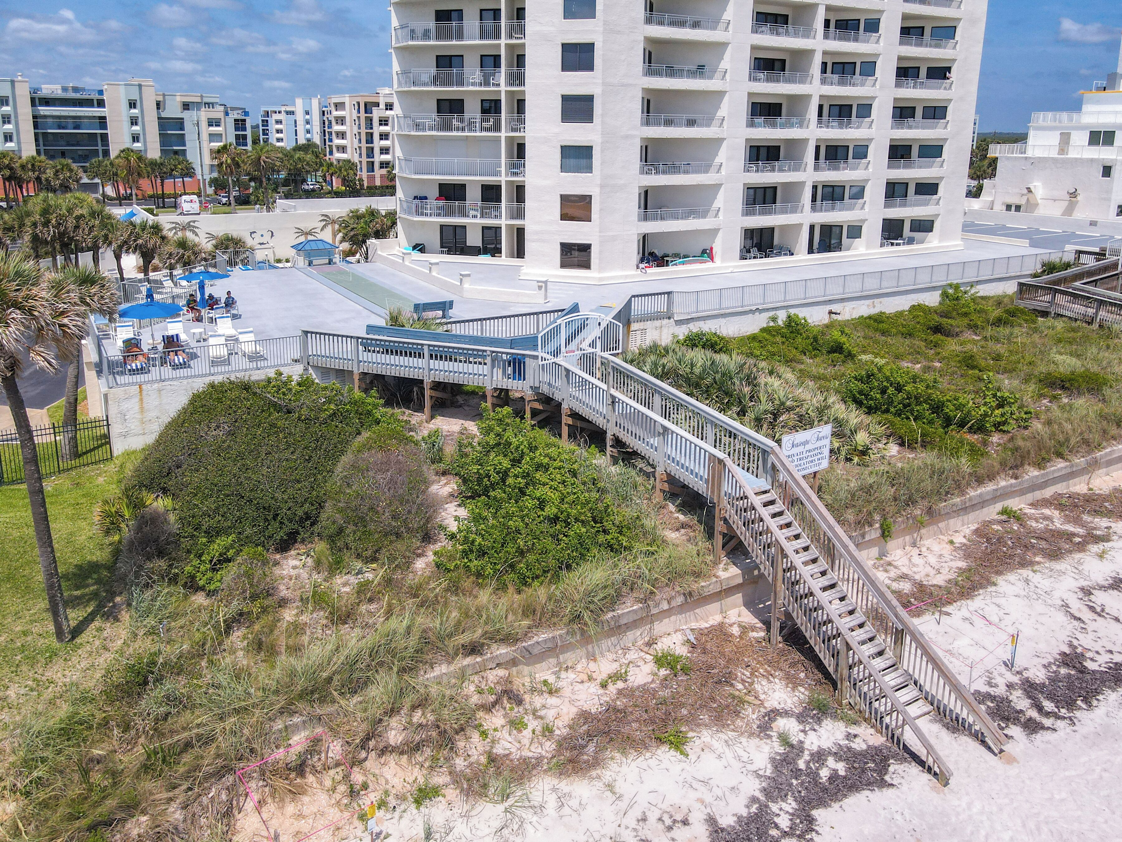 Un hotel frente a la playa con estructura de varios niveles, mobiliario exterior y una escalera que baja a la playa.