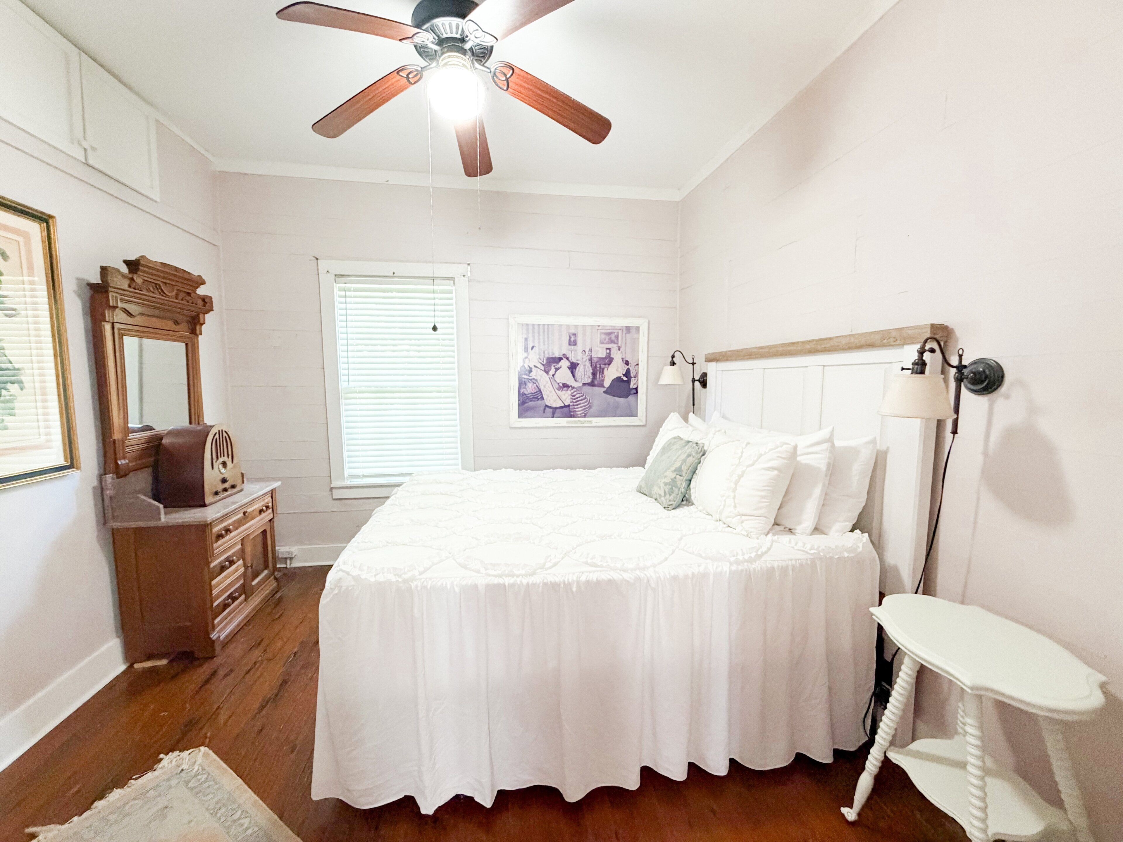 A bedroom with a bed, dresser, ceiling fan, and a small white table.
