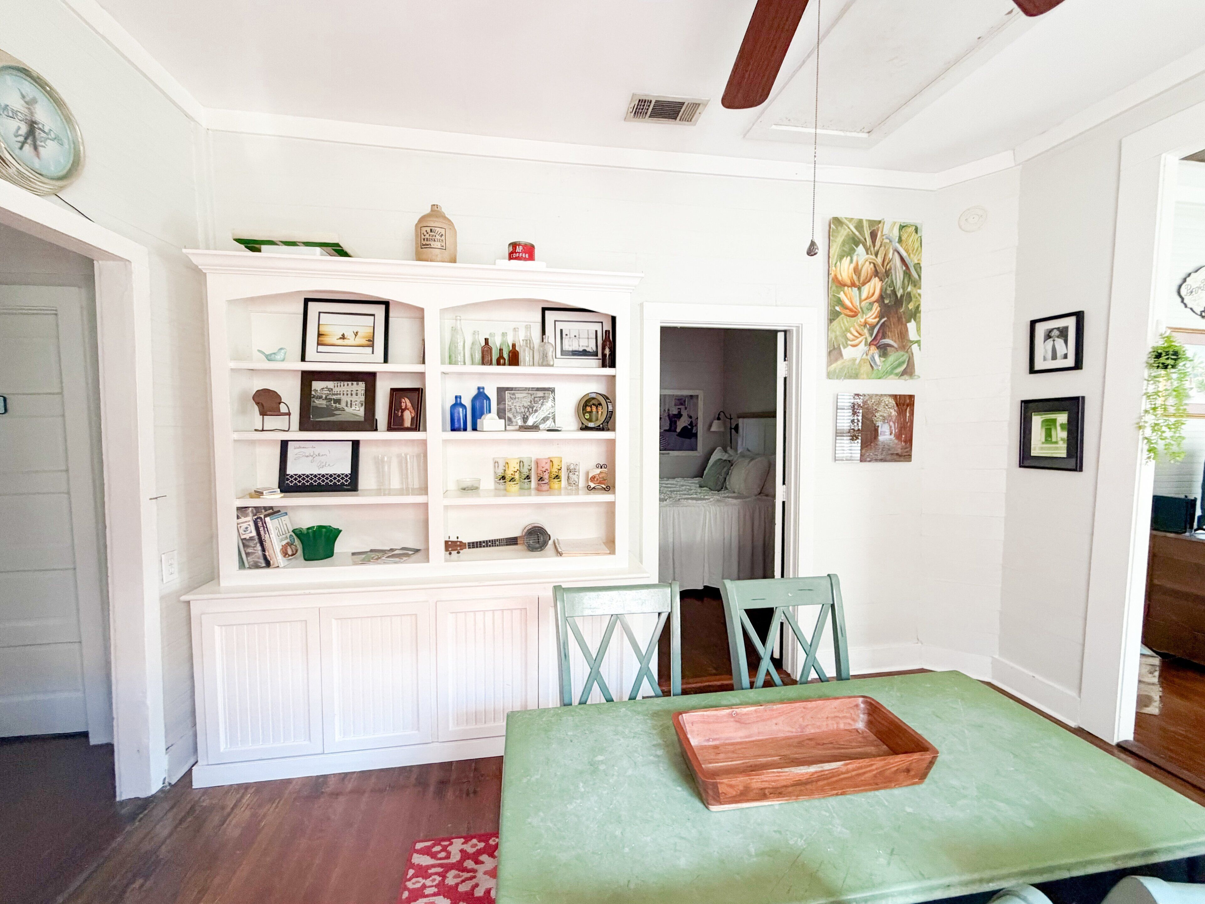 Un comedor con una mesa verde, muebles blancos con estantes y vista a un dormitorio.