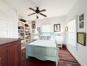 A bedroom with a bed, a ceiling fan, a bookshelf, a window with blinds, and a door.