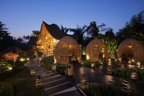 Un resort con edificios de techo de paja y estructuras en forma de cúpulas, rodeado de senderos iluminados y exuberante vegetación tropical.