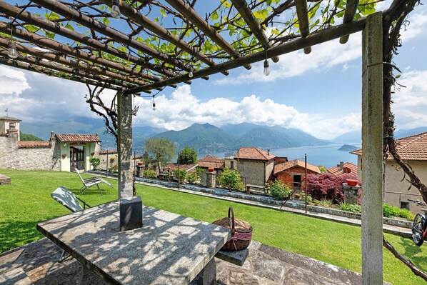 Une table en pierre avec un panier, une pergola couverte de vignes et une vue sur des maisons et des montagnes.