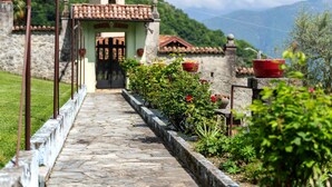 Un chemin de pierre mène à un bâtiment dont le toit est recouvert de tuiles, et qui est entouré de verdure et de pots de fleurs rouges.