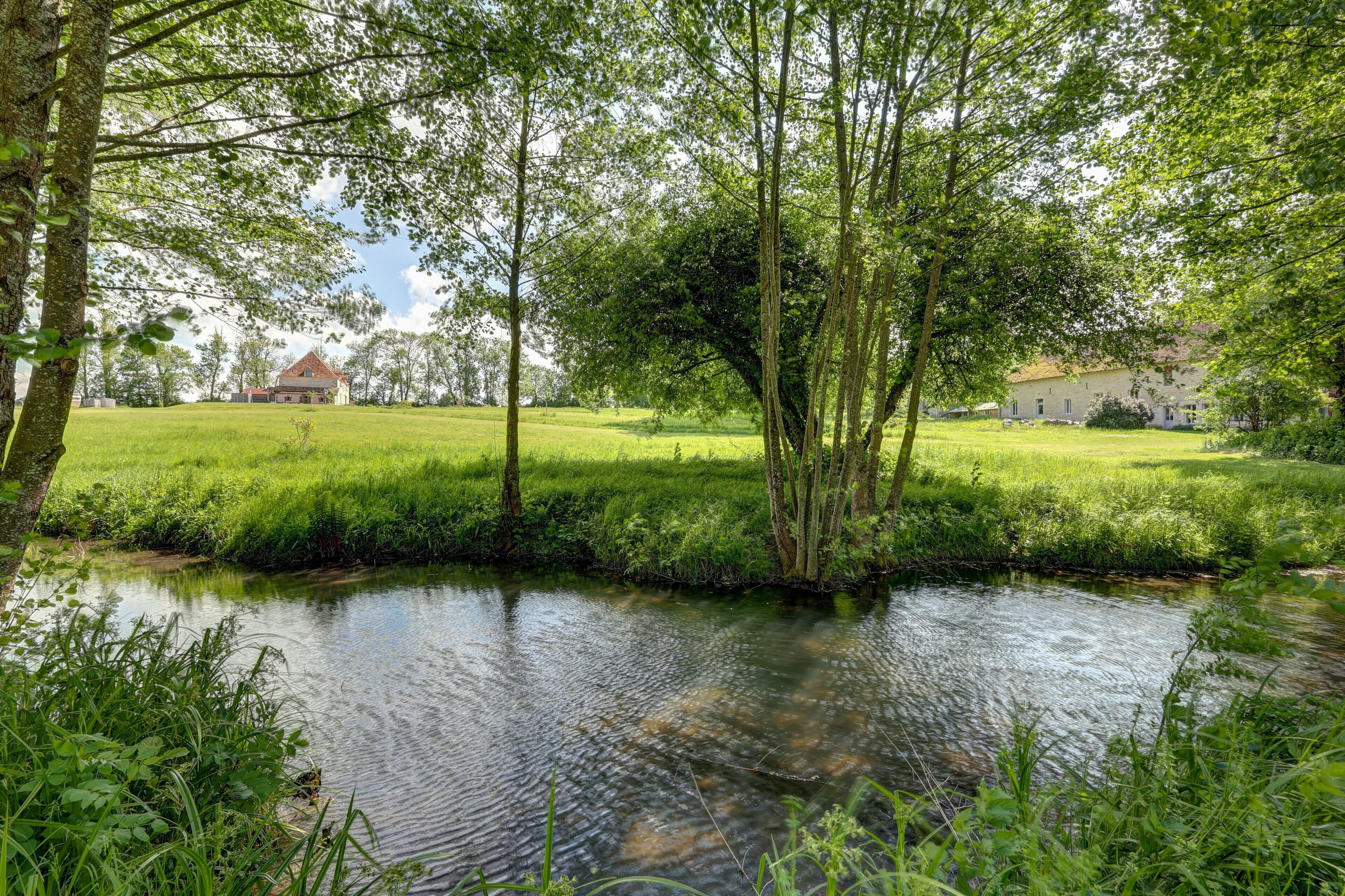 A serene riverside landscape with lush greenery and two buildings in the background.