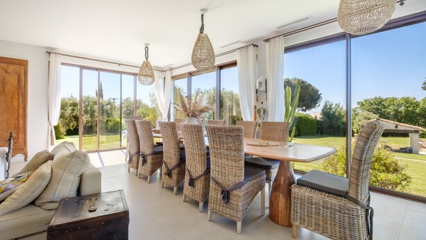 Un comedor con sillas de mimbre, una mesa de madera y grandes ventanales con vistas al exterior.