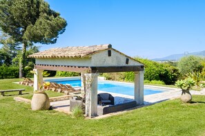 A covered outdoor seating area with a pool, surrounded by greenery and a clear sky.