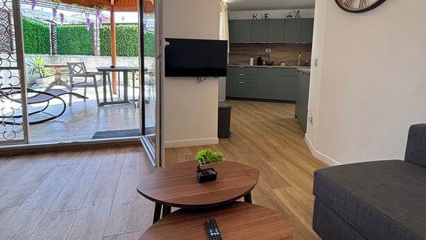 Una sala de estar moderna con un sofá, una mesa de centro con un control remoto, un televisor montado en la pared y vistas a la cocina y al patio exterior.
