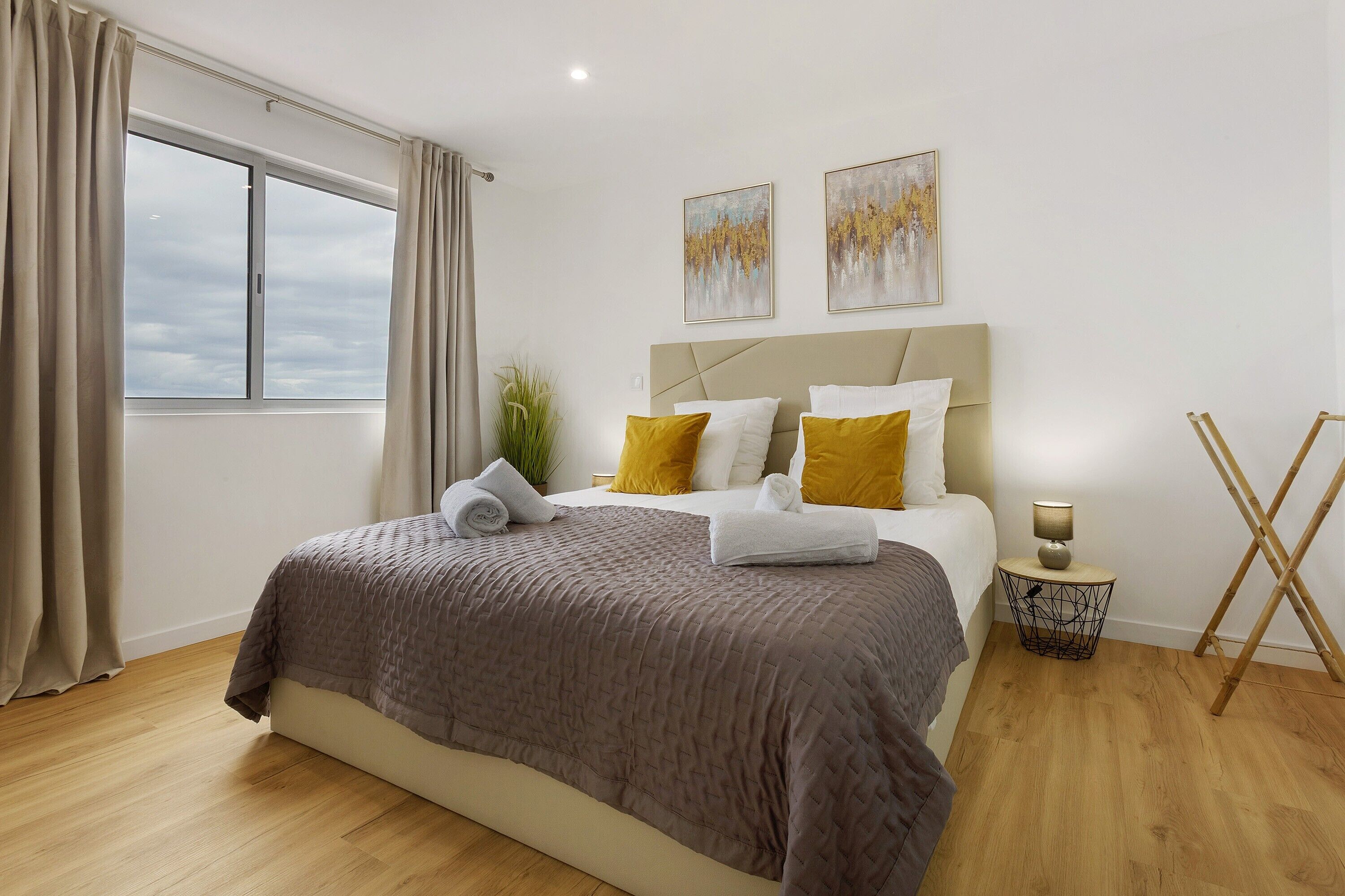 A bedroom with a bed, a window, a bedside table with a lamp, and two paintings on the wall.