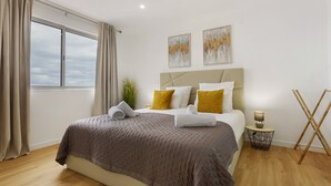 A bedroom with a bed, a window, a bedside table with a lamp, and two paintings on the wall.