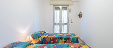 A small, brightly lit room with two beds, a window with curtains, and a wall-mounted air conditioner.
