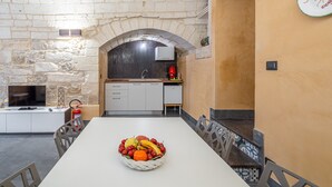 Un coin repas avec un mur en pierre, une cuisine à l’arrière-plan et un panier de fruits sur la table.