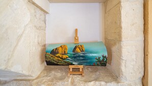 Une peinture représentant un paysage côtier avec des falaises et des cactus, un petit tabouret en bois et un mur blanc.