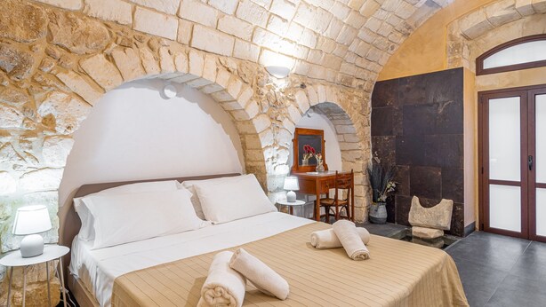 Une pièce avec un mur en pierre, un lit recouvert de draps blancs, un bureau en bois et une chaise.