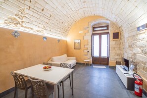 A room with a stone wall, a white sofa, a dining table with chairs, a small side table, and a fire extinguisher.