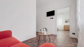 Een moderne kamer met een rode bank, een klein rond tafeltje met een waterkoker, een flatscreen televisie en een houten deur naar een andere kamer.