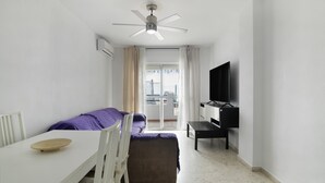A room with a bed, a TV, a ceiling fan, a table, and a window with curtains.