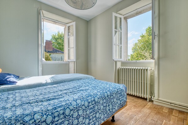 A bedroom with a bed, two windows, a radiator, and a view of the outside.