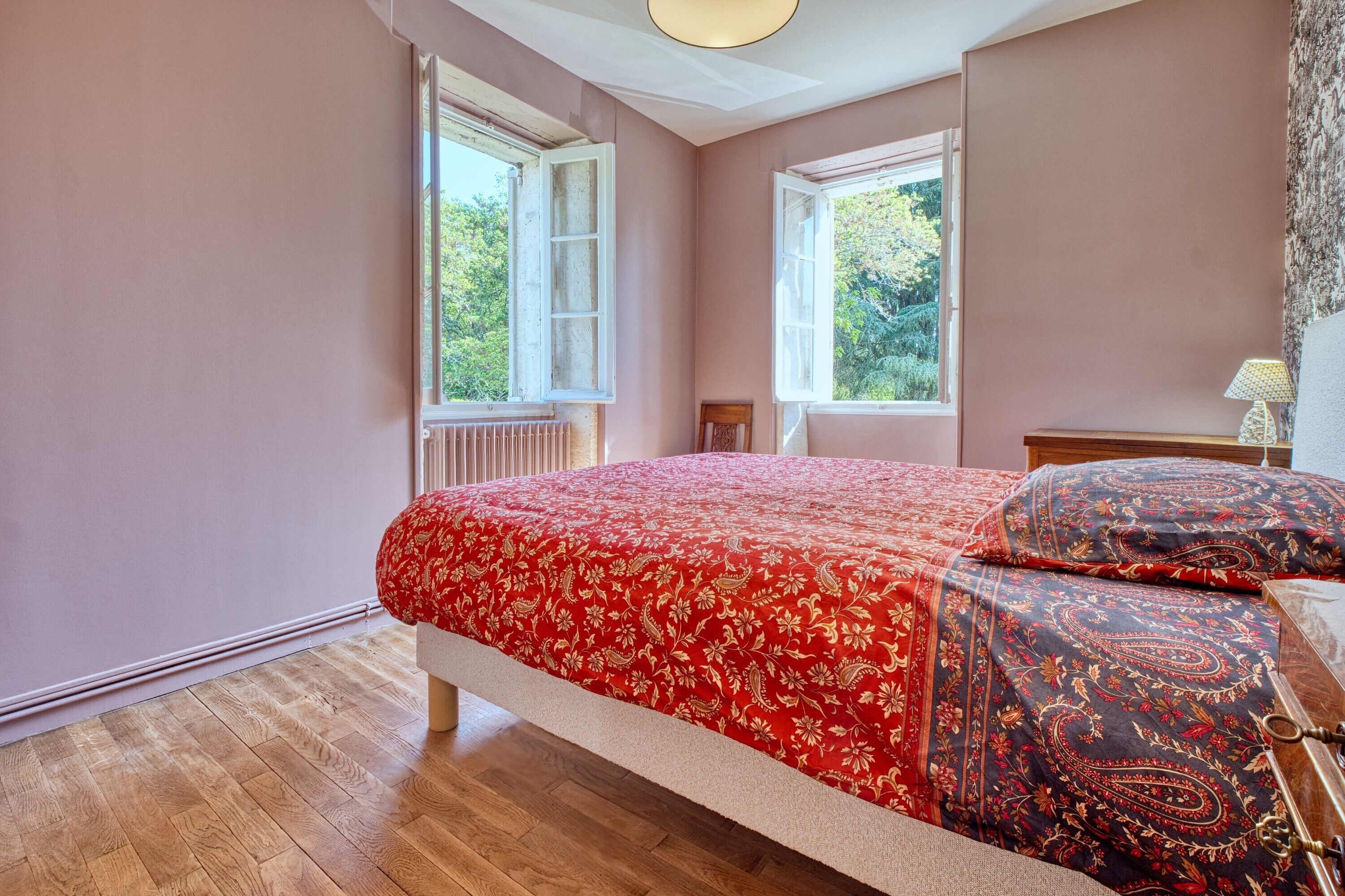 Un dormitorio con cama, dos ventanas, un radiador y una lámpara.