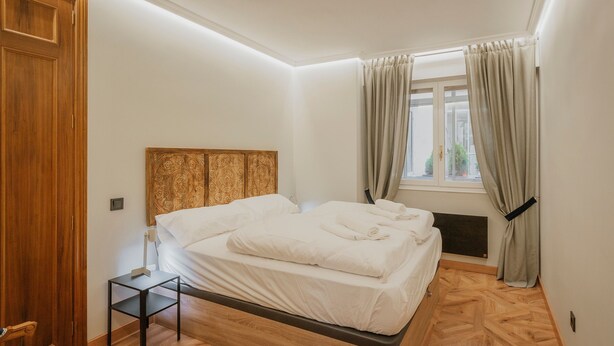 A bedroom with a wooden headboard, a bed with white linens, a small bedside table, and a window with curtains.