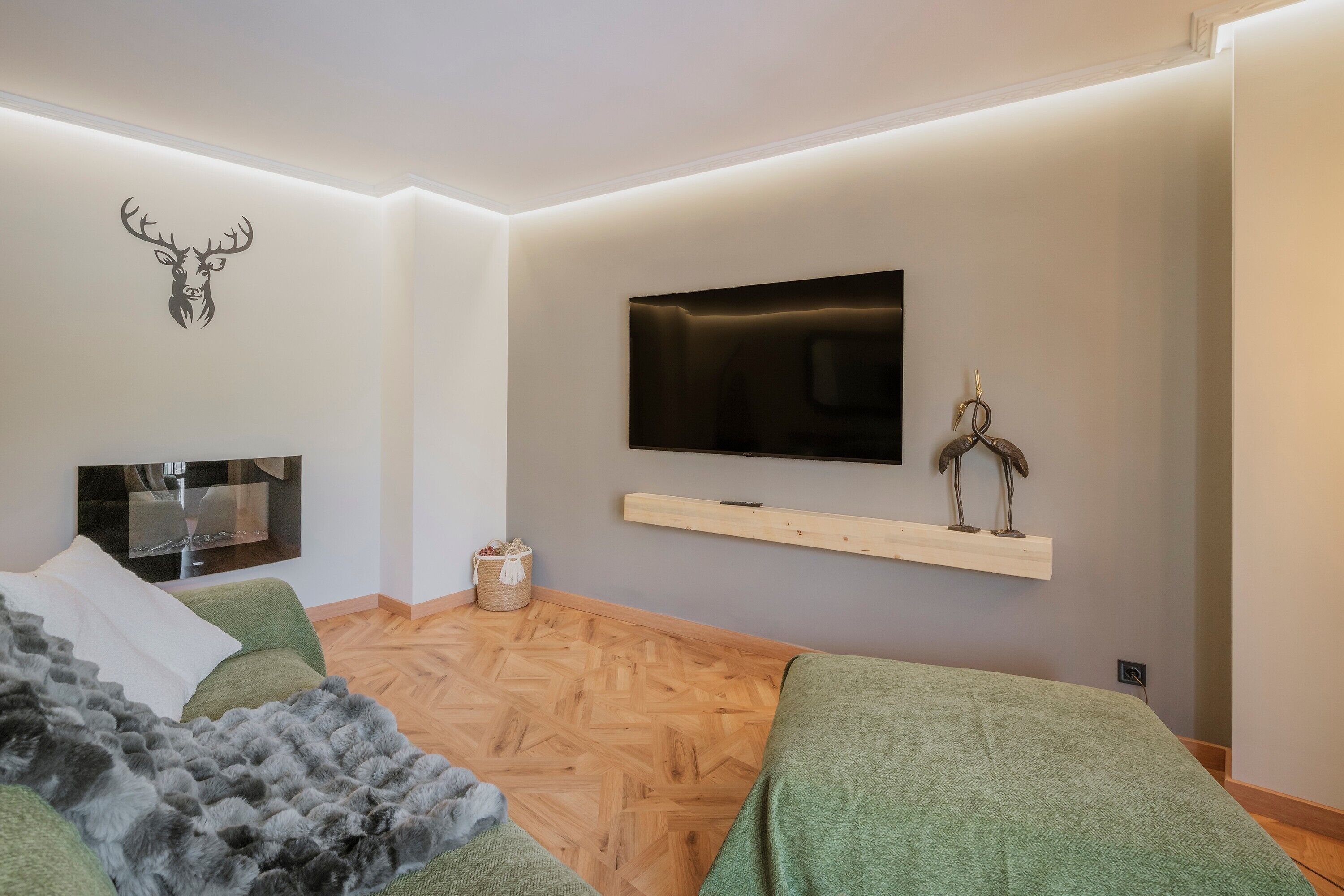 A modern bedroom with a wooden floor, a mounted TV, a mounted deer head decoration, and a bed with a green blanket.