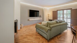 A modern living room with a sofa, a TV mounted on the wall, a window with blinds, and a dining area with a table and chairs.