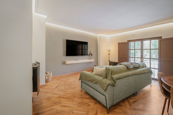 Un salon moderne comprenant un canapé, un téléviseur fixé au mur, une fenêtre avec des stores et un coin repas avec une table et des chaises.